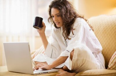Women working two jobs - woman working from home on sofa holding coffee on laptop